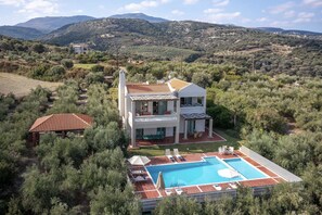 A two-story villa with a swimming pool, surrounded by dense vegetation and mountains in the background.