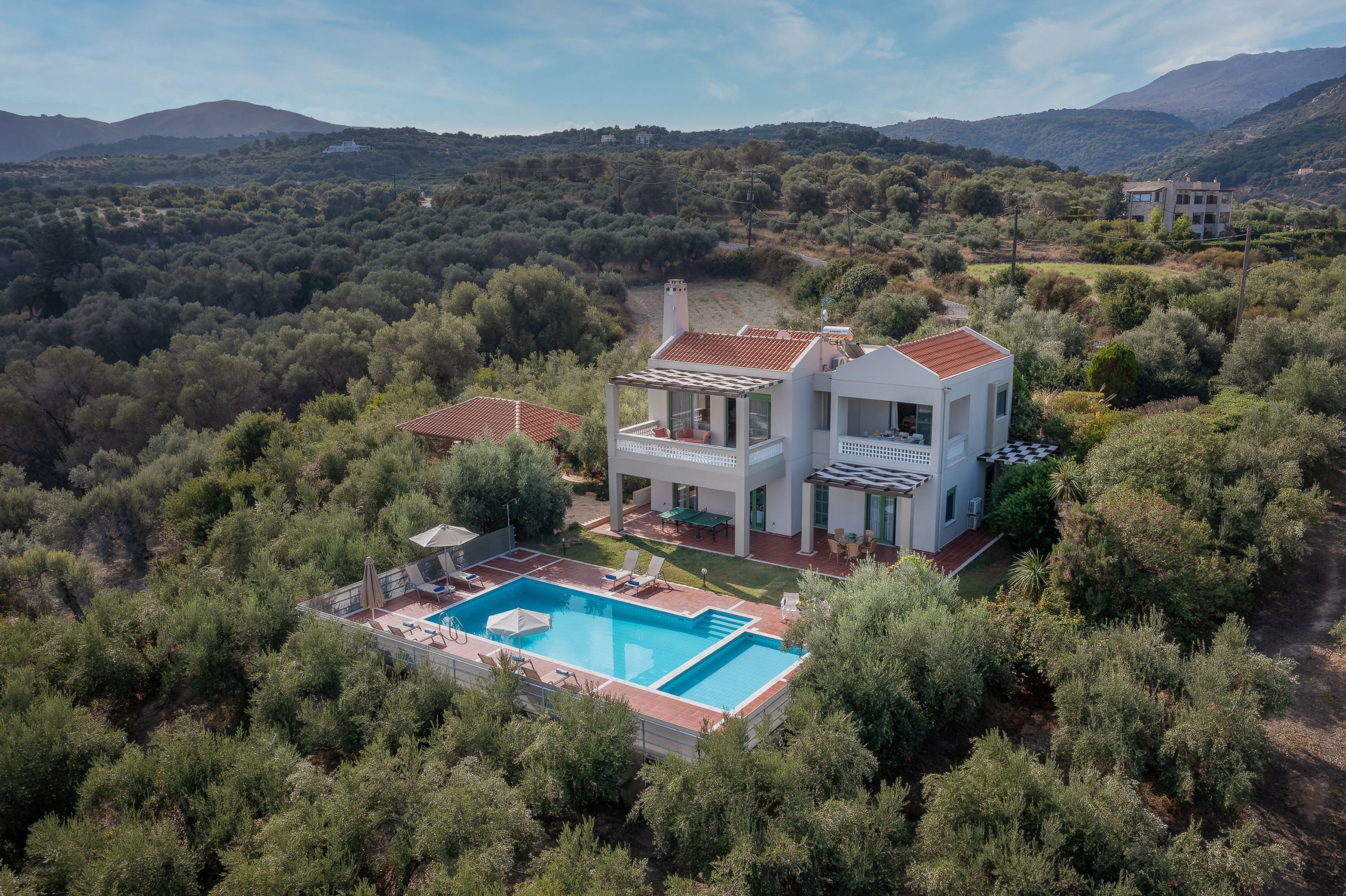 Uma casa moderna de dois andares com piscina, rodeada por vegetação densa e colinas.