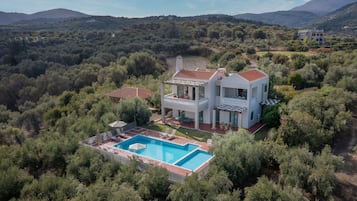 Uma casa moderna de dois andares com piscina, rodeada por vegetação densa e colinas.