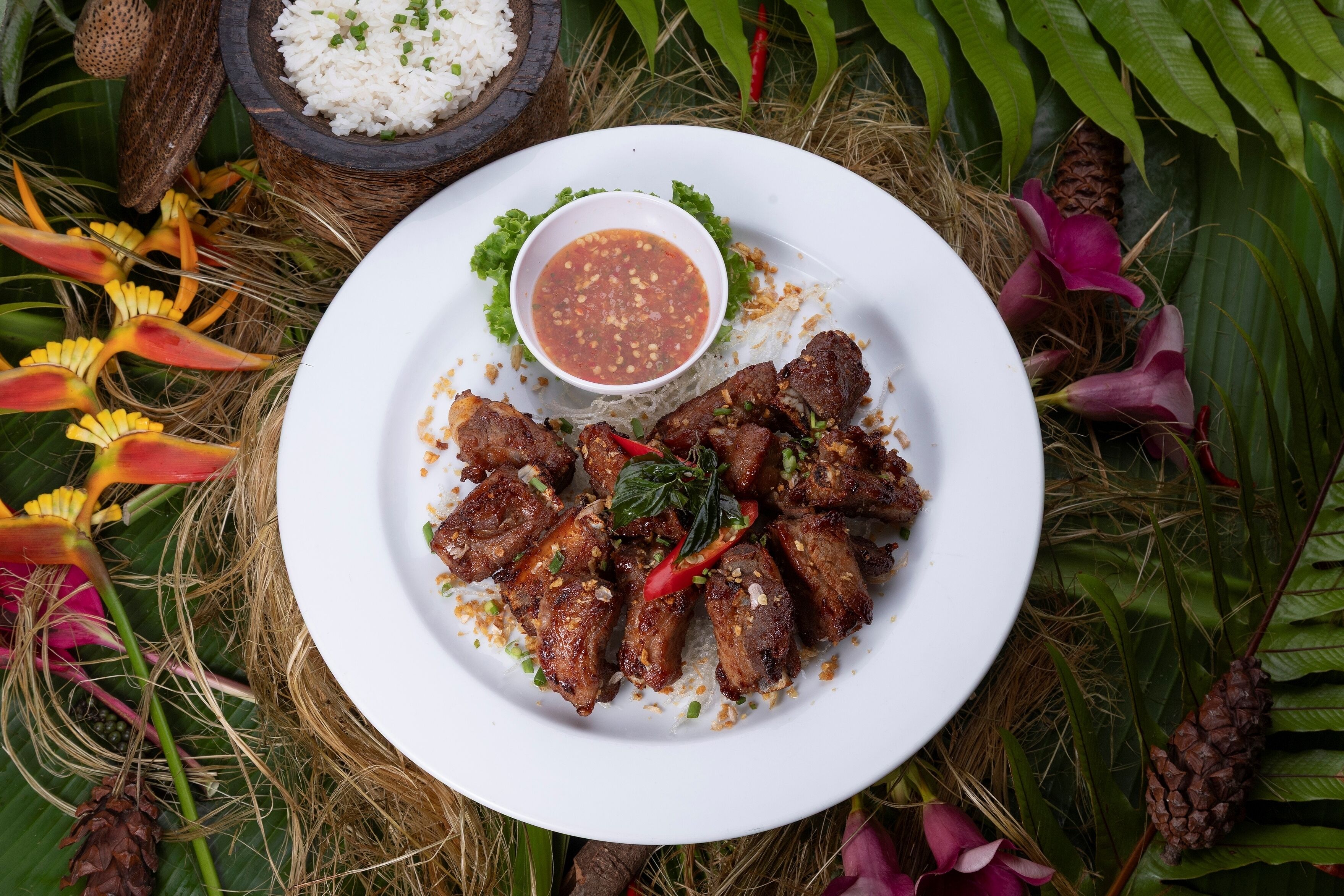 A plate of grilled meat with a side of rice and a dipping sauce, surrounded by natural elements and flowers.