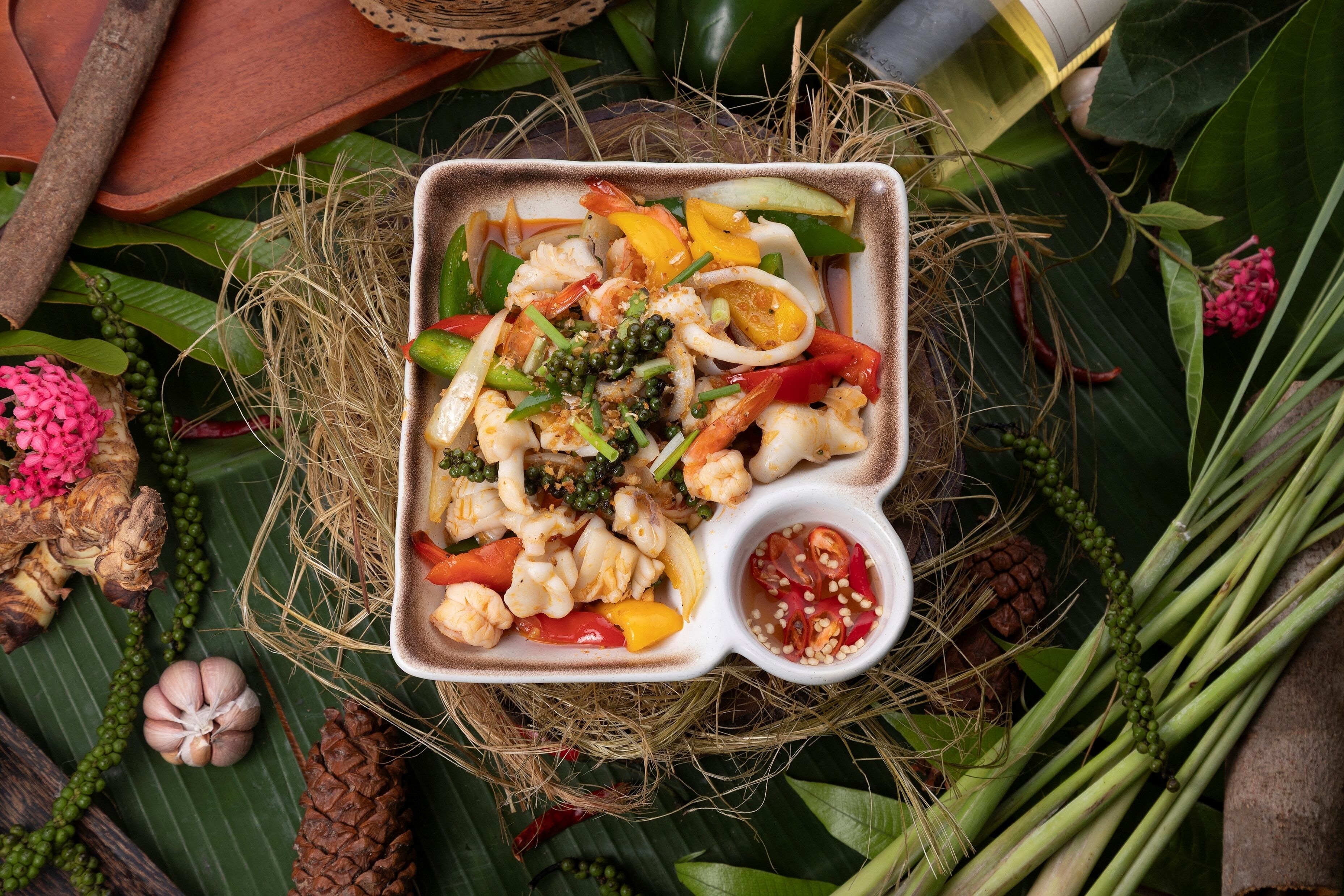 A square dish with various vegetables and seafood, surrounded by natural elements like leaves and a small bowl of sauce.