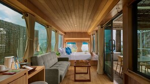 Una habitación moderna con techo de madera, un sofá, una cama con almohadas azules, una mesita y ventanas amplias con vistas a la vegetación.
