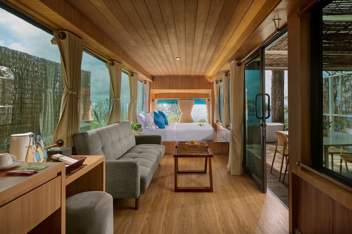 Una habitación moderna con techo de madera, un sofá, una cama con almohadas azules, una mesita y ventanas amplias con vistas a la vegetación.