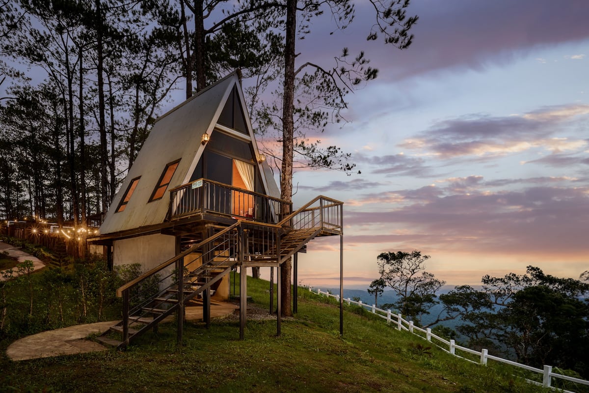 Una casa en árbol con una escalera que lleva a un balcón, rodeada de árboles y una valla blanca.