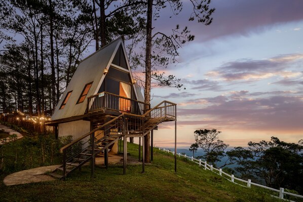 Una casa en árbol con una escalera que lleva a un balcón, rodeada de árboles y una valla blanca.