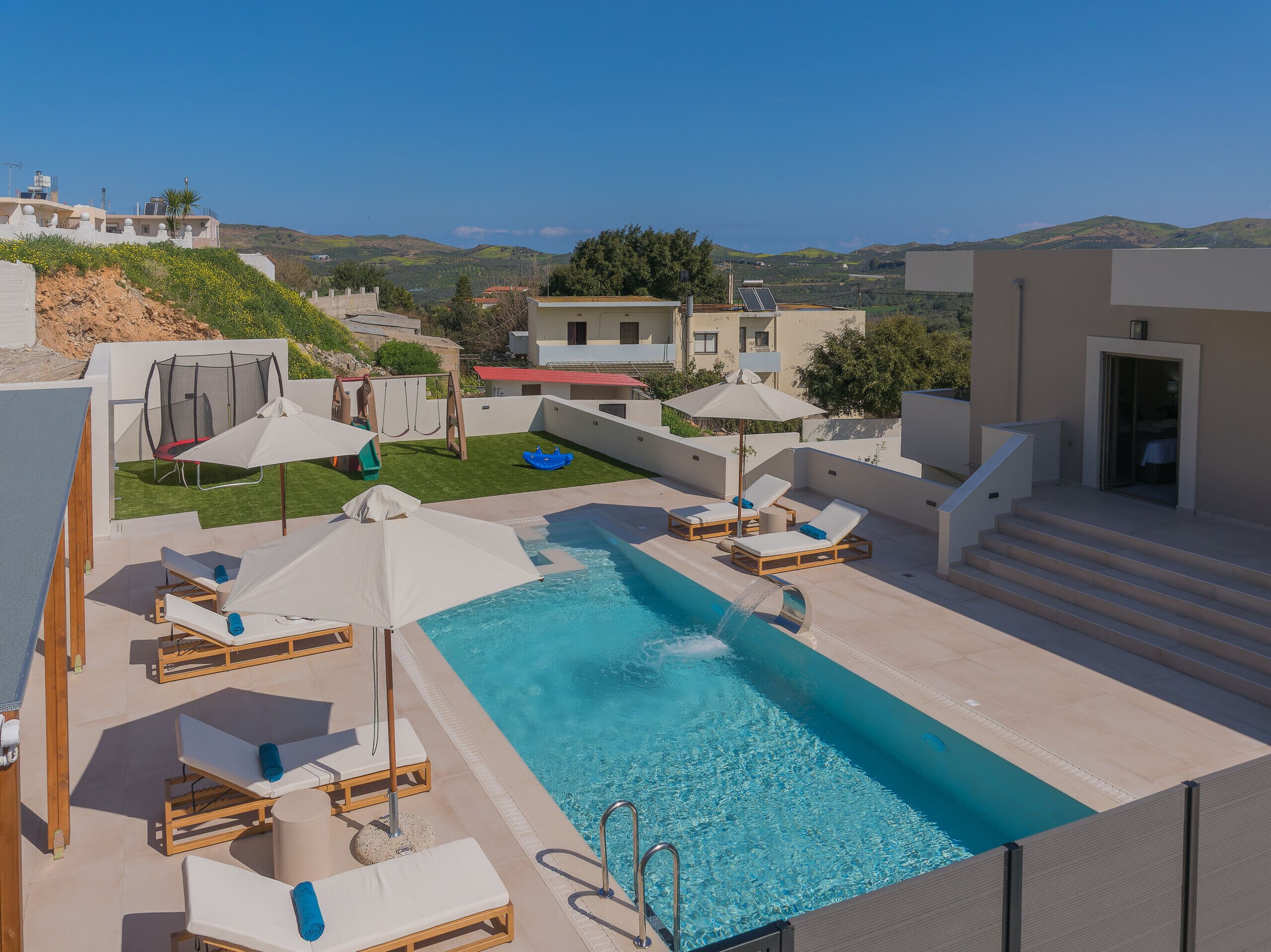 Un área de piscina en la azotea con sillas de descanso y un tobogán pequeño, rodeada de edificios y con vistas a las colinas.