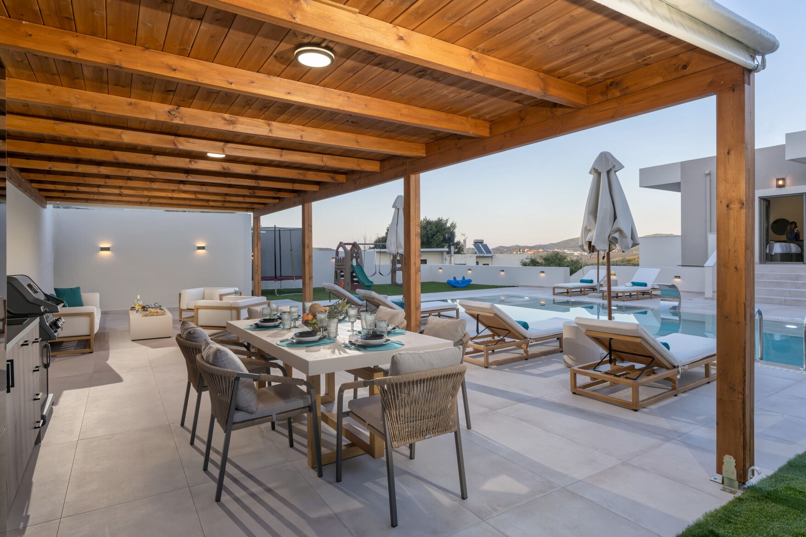 Un área de comedor al aire libre techada con piscina y sillones de descanso, que cuenta con techo de madera y paredes blancas.