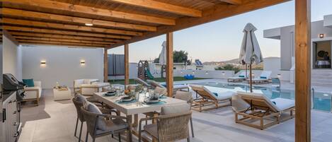 Un área de comedor al aire libre techada con piscina y sillones de descanso, que cuenta con techo de madera y paredes blancas.
