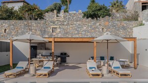 A poolside area with lounge chairs and umbrellas, a stone wall, and a kitchen area. - Elysian Villa 2 - With Private Pool (Mylopotamos)