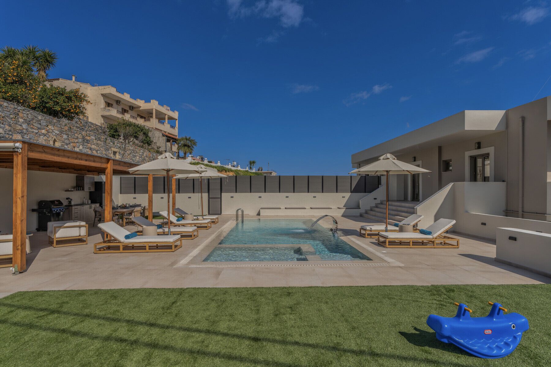 A pool area with lounge chairs and umbrellas, a small children's play area, and a stone wall with plants.