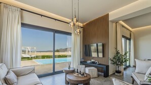 A modern living room with a large window, a flat-screen TV, and a pool view. - Villa Lux Solis - With 2 Pools (Mylopotamos)