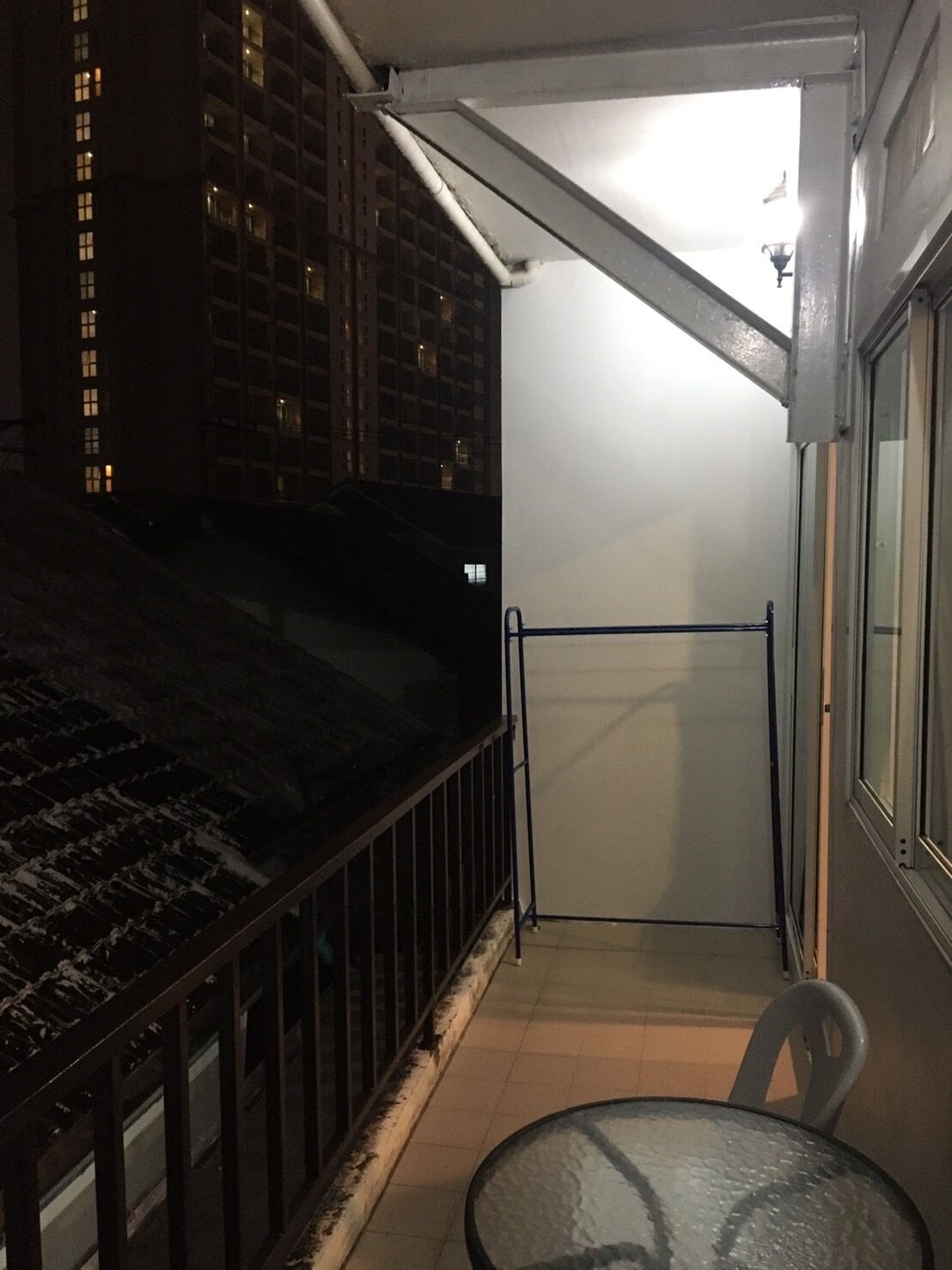Un balcon avec une table et des chaises en verre, une rambarde en métal et une vue sur un immeuble la nuit.