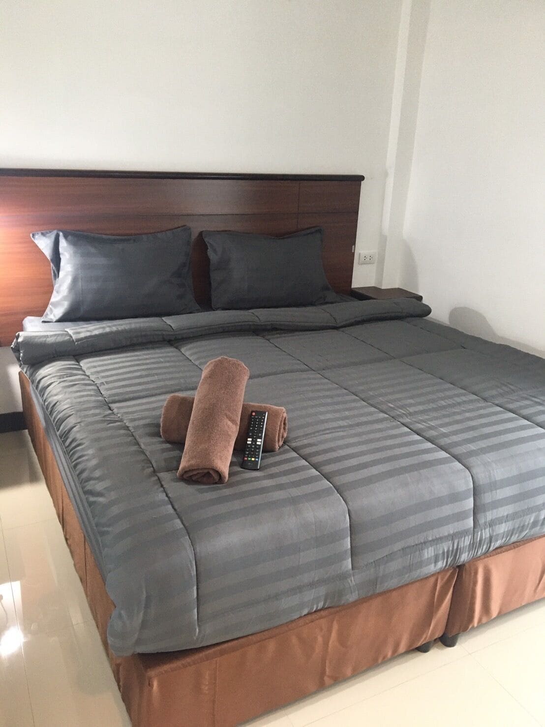 Un lit bien fait, avec une literie grise et une serviette marron, une télécommande et une tête de lit en bois.
