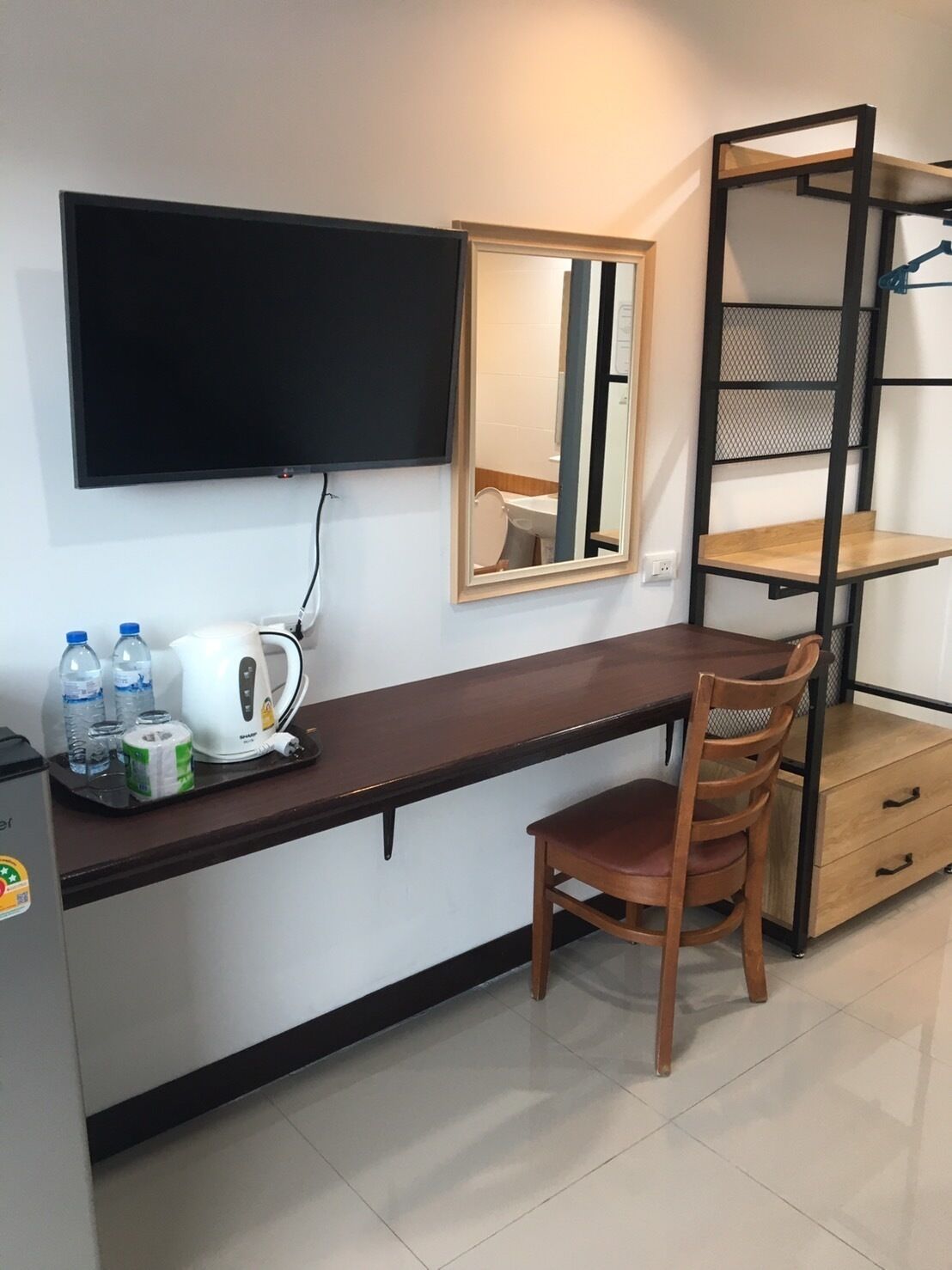 A compact hotel room with a dark wooden desk, a single wooden chair, a small kitchenette area with a kettle, and a mirror.