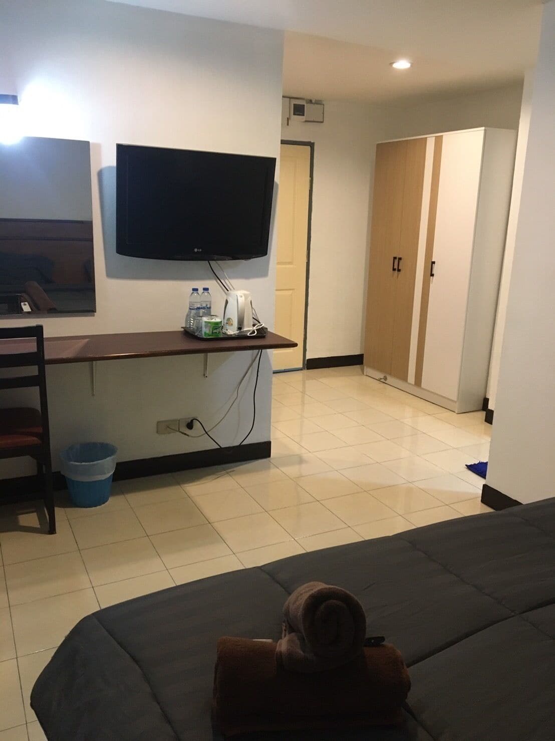 A hotel room with a bed, a desk with a flat-screen TV, a chair, a blue trash bin, and a wardrobe.