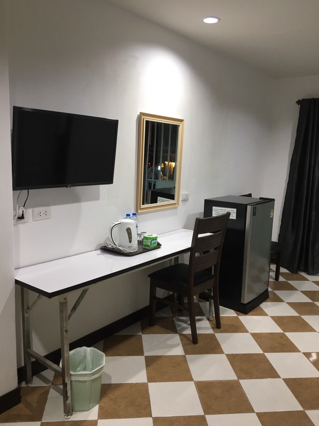 Una habitación pequeña y funcional con un escritorio blanco, una silla, un televisor montado en la pared, un espejo, un refrigerador y un cubo de basura.