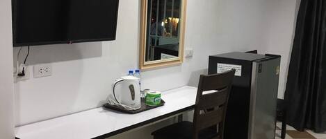 Una habitación pequeña y funcional con un escritorio blanco, una silla, un televisor montado en la pared, un espejo, un refrigerador y un cubo de basura.