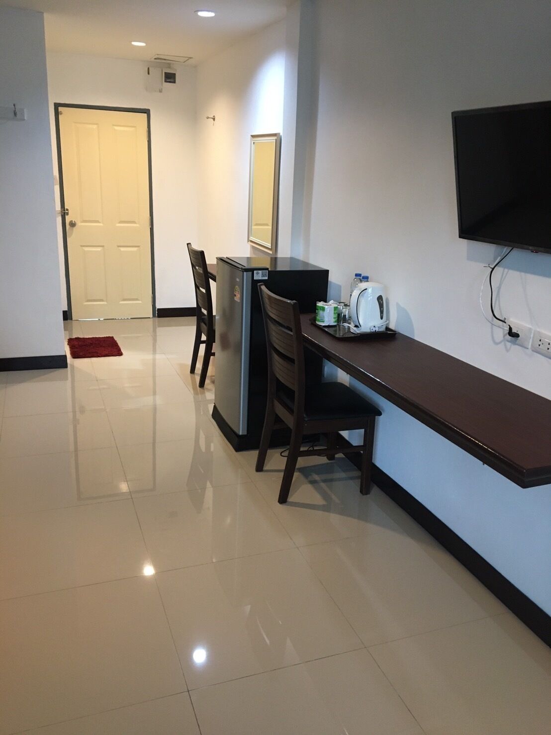 A modern kitchen with a refrigerator, a microwave, and a toaster on the counter. There is a dining table with chairs and a red rug on the floor. A TV is mounted on the wall.