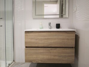 A bathroom with a white tiled wall, a wooden vanity with a mirror above it, and a glass shower enclosure. - Modern, functional studio 15 min from center with Wi-Fi and air conditioning (Málaga)