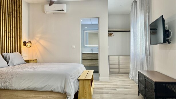 A modern bedroom with a bed, a wooden bench, a ceiling fan, an air conditioning unit, a TV mounted on the wall, and a view into another room. - Modern, functional studio 15 min from center with Wi-Fi and air conditioning (Málaga)