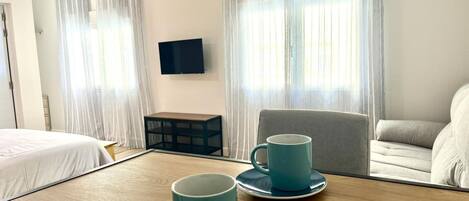 A hotel room with a bed, a wooden table with two blue mugs, a grey sofa, and a TV mounted on the wall.
