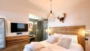 Een slaapkamer met een houten hoofdbord, een aan de muur bevestigde televisie, een bed met witte lakens en een doucheruimte zichtbaar door een glazen deur.