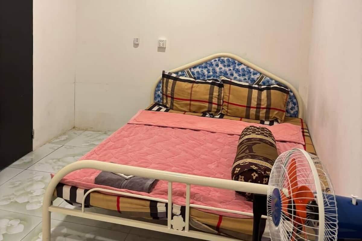 A small, single-bed room with a white metal frame, a pink quilted bedspread, and a floral-patterned headboard. A standing fan is positioned to the right of the bed.