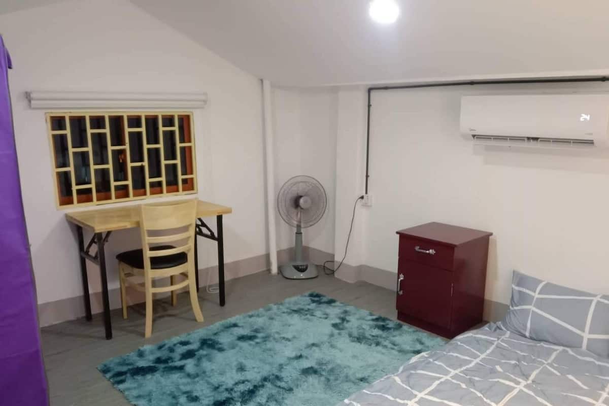 A small, clean room with a bed, a desk with a chair, a fan, and a wardrobe.