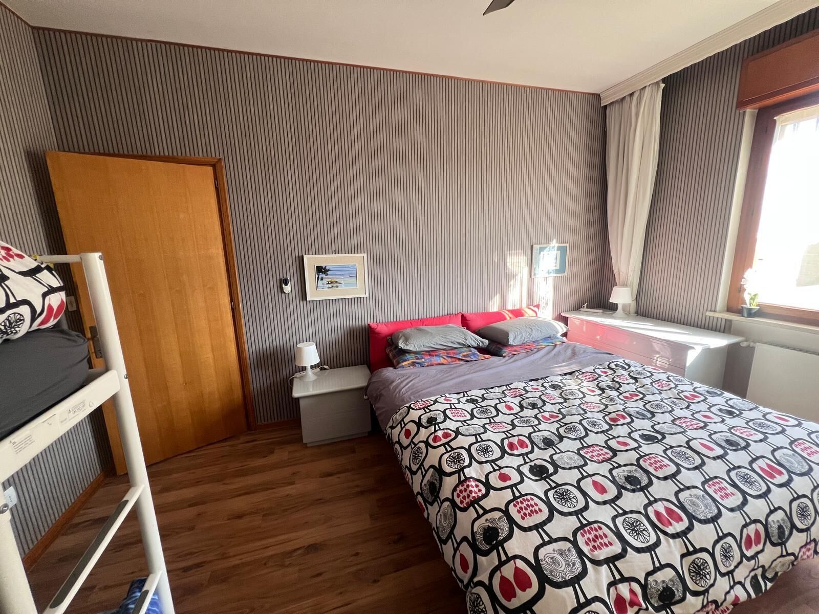 A bedroom with a bunk bed, a bedside table with a lamp, a bed with patterned bedding, and a window with curtains.
