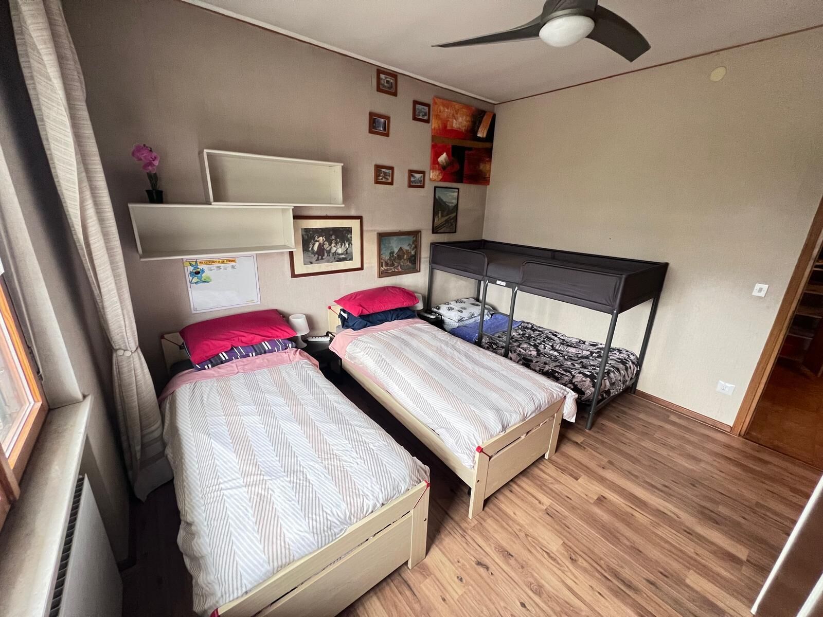Quarto com duas camas de solteiro, um ventilador de teto, uma estante com quadros e piso de madeira.