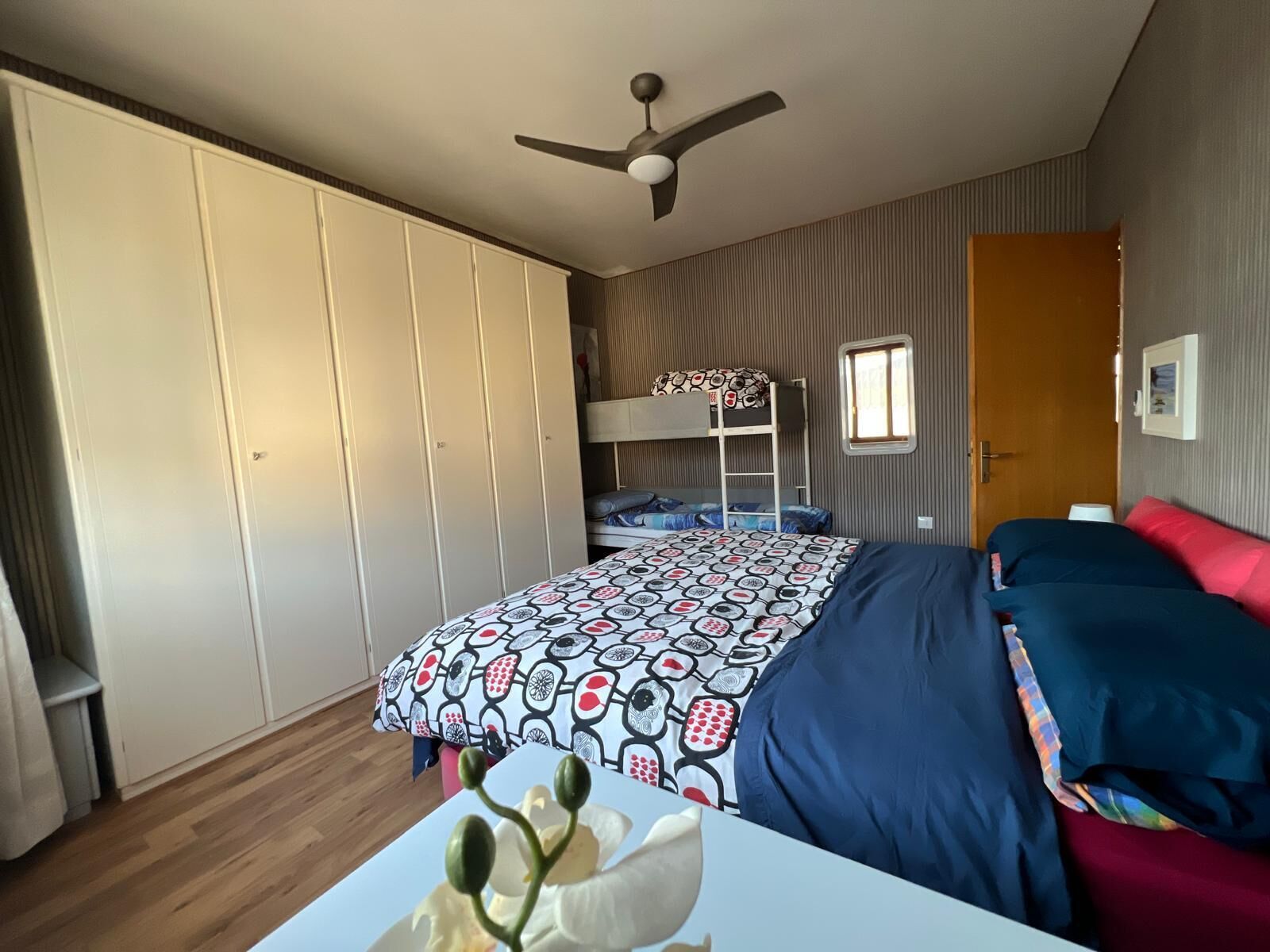 Un dormitorio con una cama litera, un armario, un ventilador de techo y una ventana.