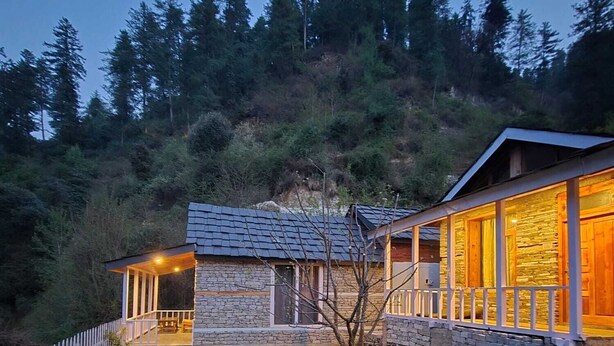 A stone building with a porch, a fenced yard, and a forested hillside in the background. - Jibhi Village (Banjar)