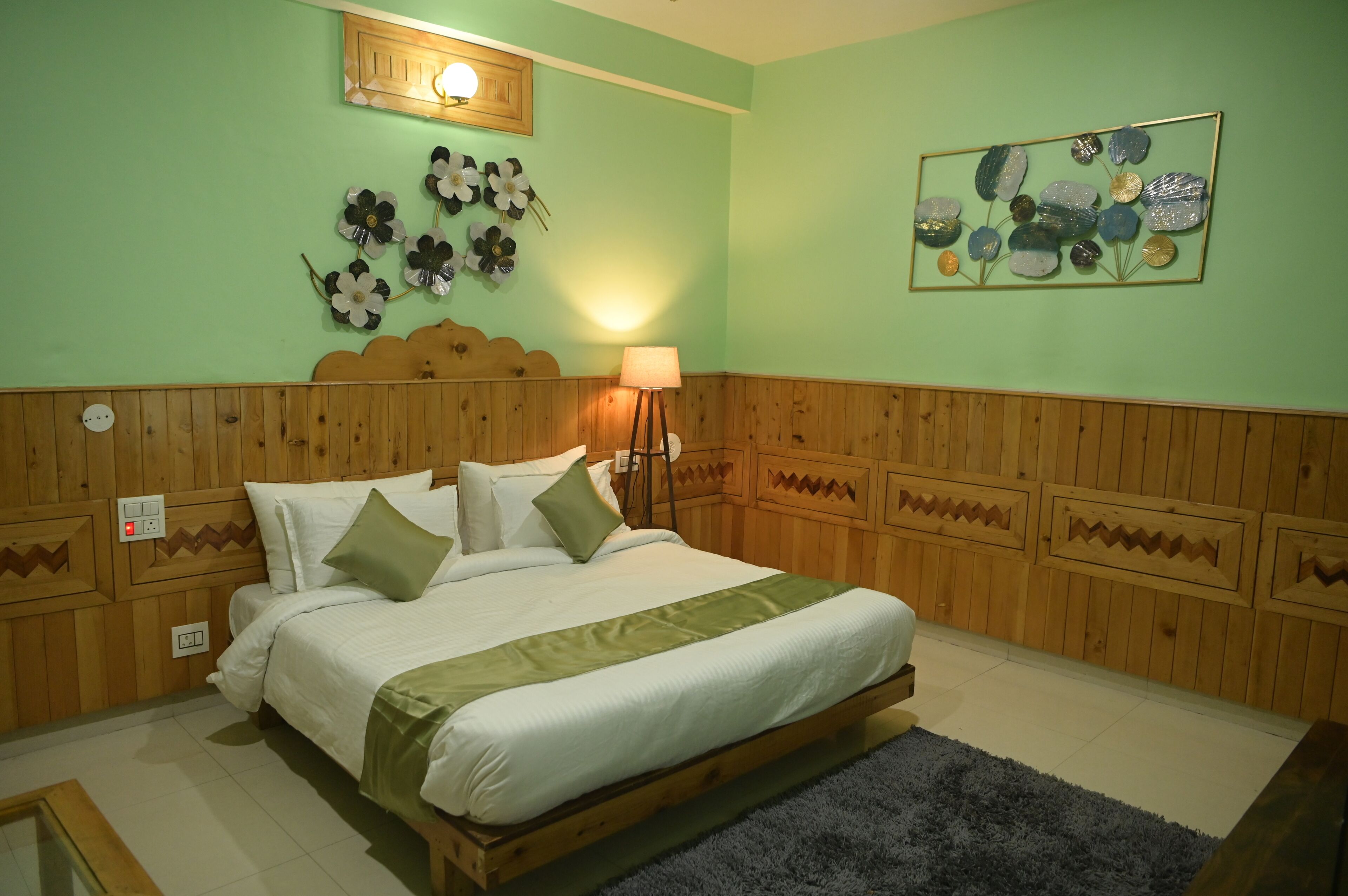 Un dormitorio con cabecera de madera, cama con ropa de cama blanca y detalles verdes, una lámpara de pared y una obra de arte floral.