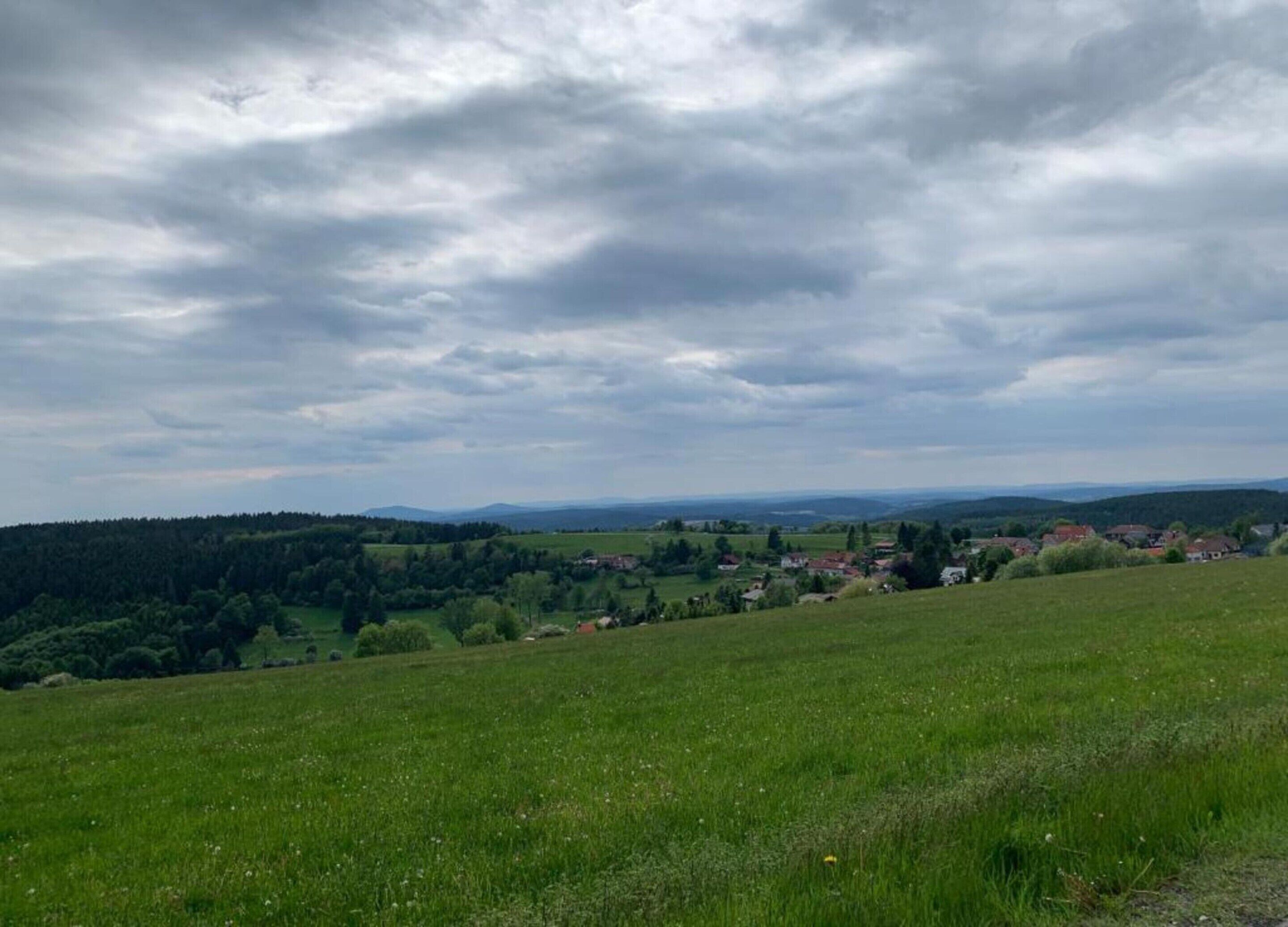 A rural landscape with rolling hills, a village, and a forested area under a cloudy sky.