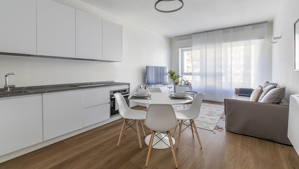 A modern kitchen with white cabinets, a dining area with a round table and chairs, and a grey sofa in a bright living space with large windows. - Le Barche66 Venice Apartments (Mestre)