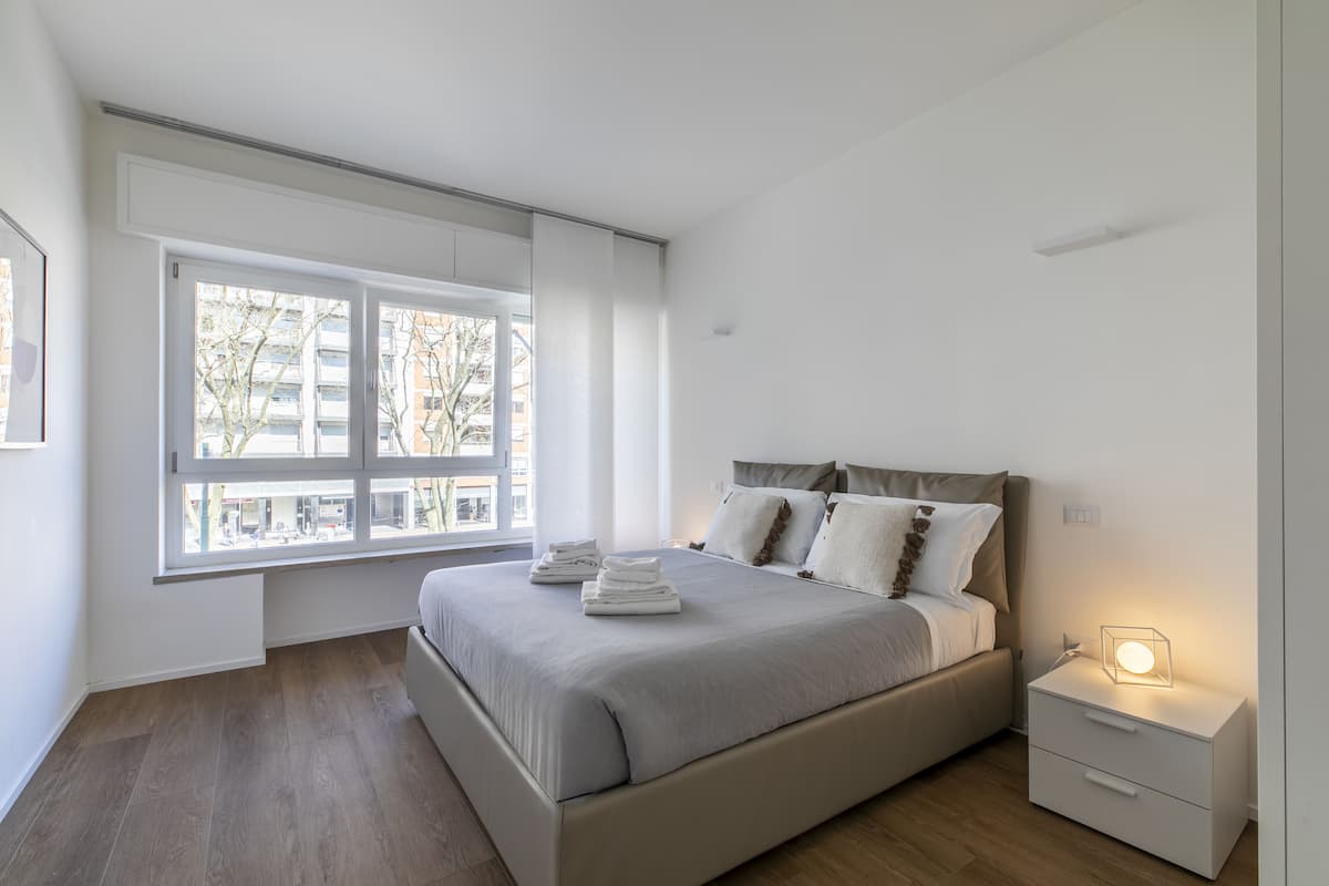 A modern bedroom with a large window, a bed with neatly folded towels, a bedside table with a lamp, and a nightstand.