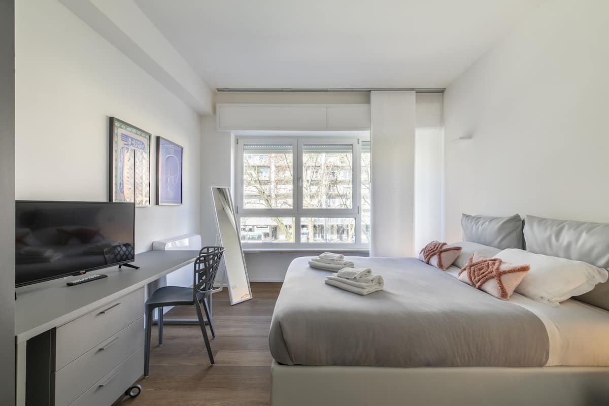 A modern hotel room with a large bed, a desk with a TV, and a window with a view of buildings.