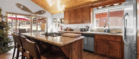 A kitchen with wooden cabinets, a central island with bar stools, a stainless steel refrigerator, and a window with a view of the outside.