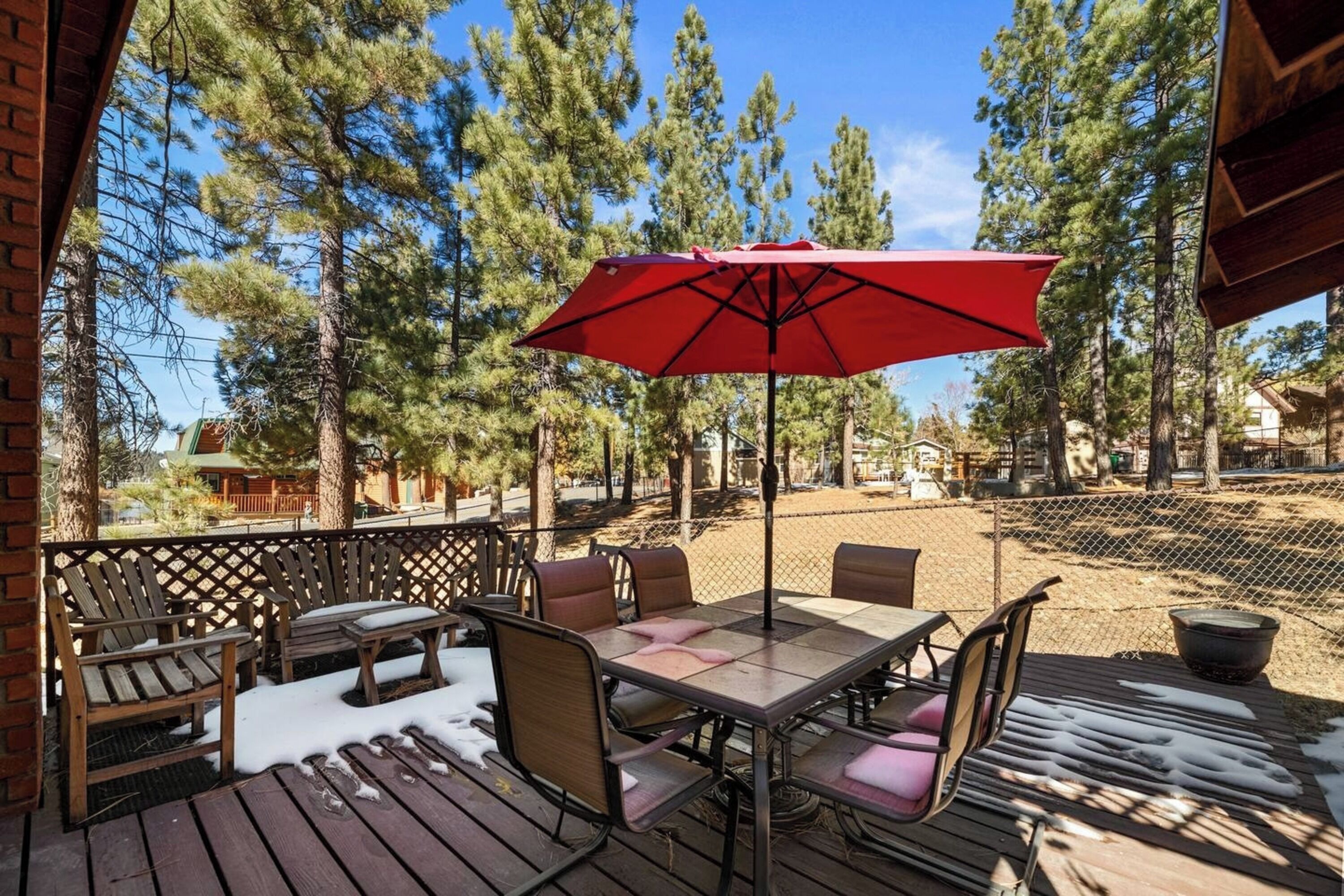 An outdoor patio with wooden furniture, a red umbrella, and a view of tall pine trees.