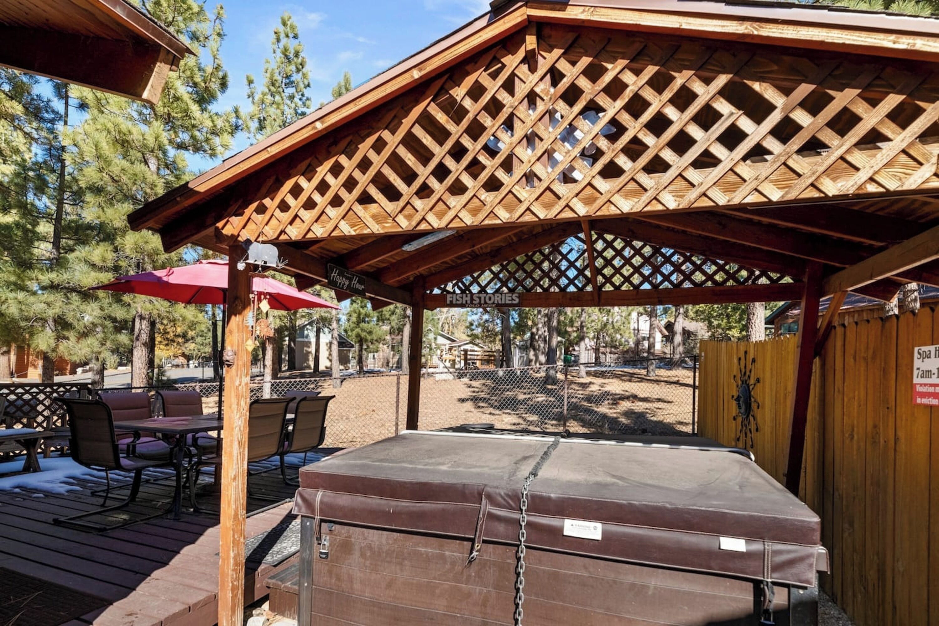 A covered outdoor seating area with a wooden lattice roof, a hot tub, and a patio table with chairs.