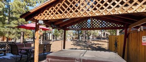 A covered outdoor seating area with a wooden lattice roof, a hot tub, and a patio table with chairs.
