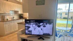 Een woonkamer met een televisie waarop een muziekvideo draait, een keukenhoek en uitzicht op een zwembad en een huis.