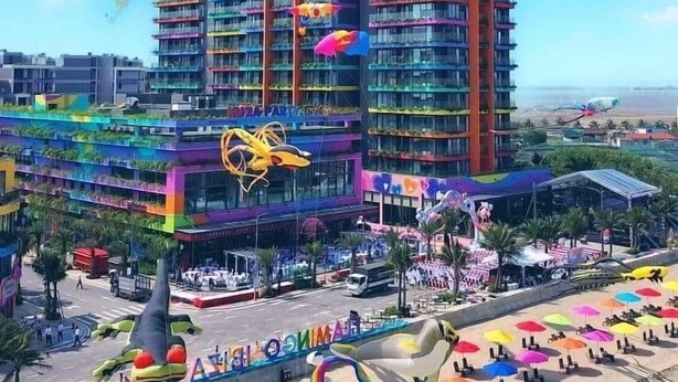 Un hotel de varios pisos con balcones, una zona de playa con sombrillas y un cielo despejado.
