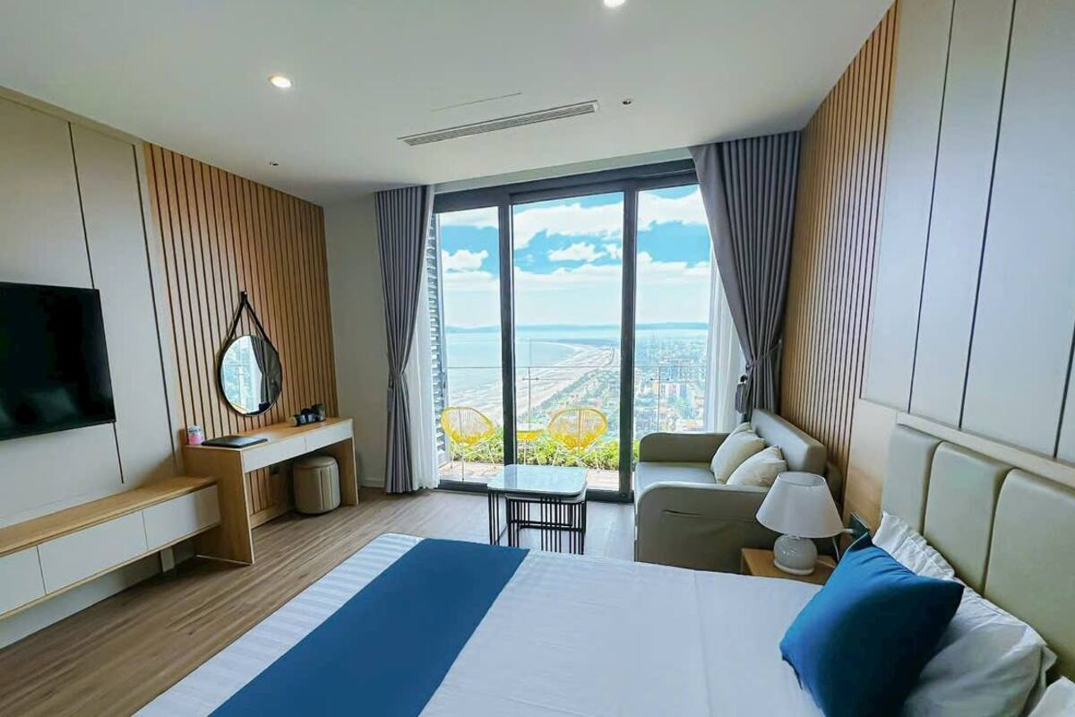 Quarto de hotel moderno com uma cama grande, uma mesa, uma cadeira, uma televisão e vista para a cidade.