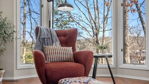 A red armchair with a patterned throw and pillows, positioned in front of a large window with a view of trees and a clear sky. - Treetop Trailhaven-Direct Access to MTB Trails! (Bella Vista)