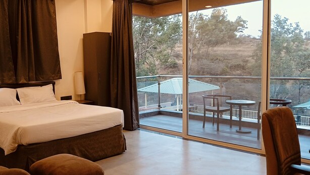 A hotel room with a bed, a nightstand, a lamp, a large window with a view of the outdoors, and a balcony with outdoor furniture. - Mountain Meadows Resort (Mahabaleshwar)