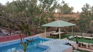 A swimming pool with a green umbrella, outdoor seating, and a view of trees and hills. - Mountain Meadows Resort (Mahabaleshwar)