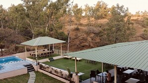 A pool with a pergola and outdoor seating area, surrounded by a landscaped garden and a hilly backdrop. - Mountain Meadows Resort (Mahabaleshwar)