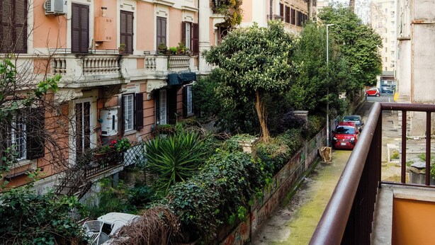 A narrow urban street with buildings on both sides, a tree, and a red car parked on the right. - Cereate Suites (Rome)