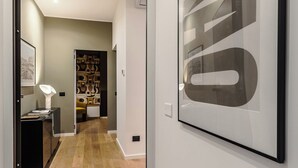 A hallway with a wooden floor, a framed artwork on the wall, and a door leading to a room with a desk and a lamp. - Cereate Suites (Rome)